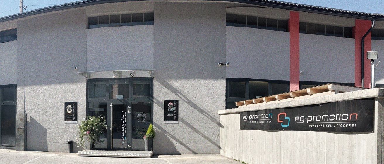 Ein graues Gebäude mit zwei Glastüren und einer Überwachungskamera über dem Eingang. Die linke Tür hat ein Schild mit der Aufschrift 'DRUCK'. Vor dem Eingang stehen Topfpflanzen.