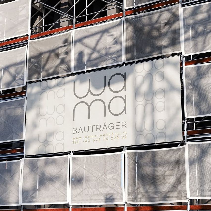 Ein großes weißes Schild mit dem Text 'WAMA' ist an einem Gebäude im Bau befestigt. Es hat eine netzartige Oberfläche und eine Website-Adresse.