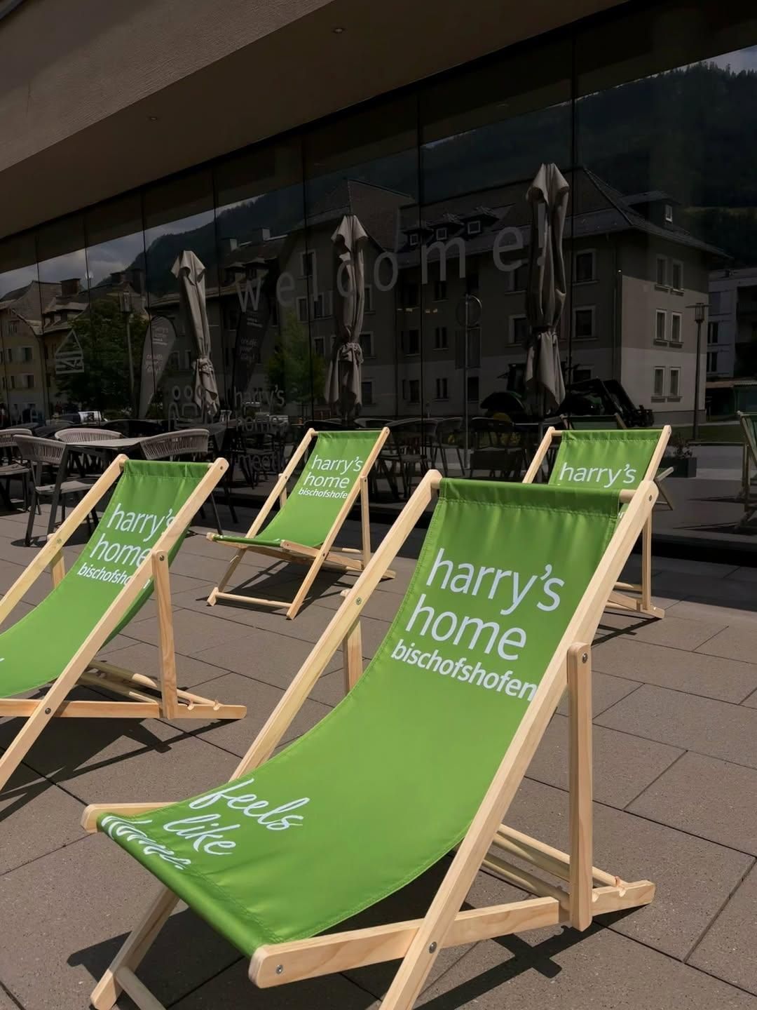 Vier grüne Klappstühle mit der Aufschrift 'Harry's Home Bischofshofen' sind ordentlich auf einer Terrasse angeordnet. Die Stühle stehen auf einem gefliesten Boden, mit Reflexionen von Gebäuden und Grünpflanzen in einer nahen Glaswand.