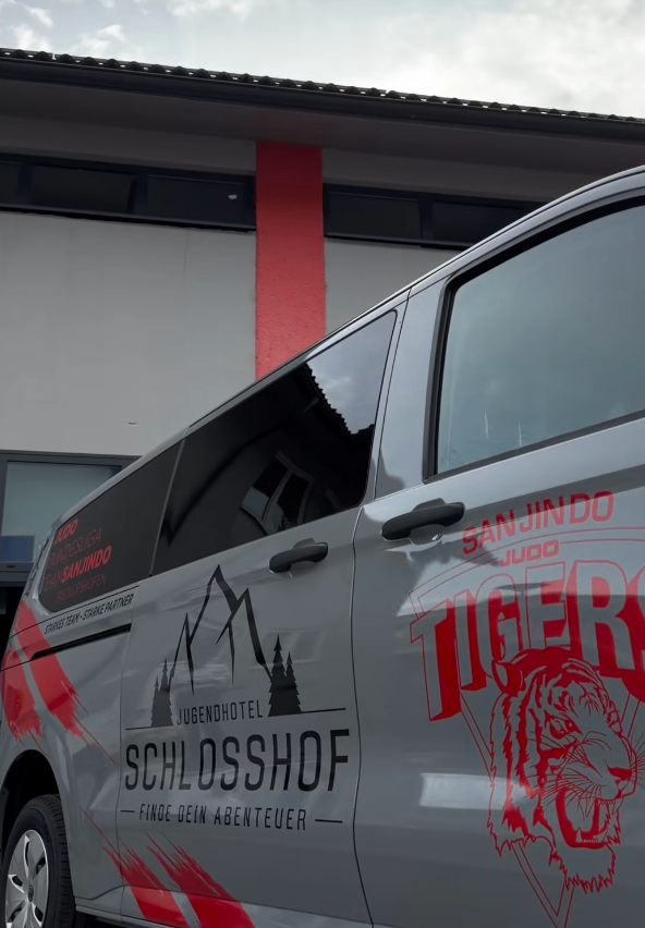Ein Van mit 'Tigers' und 'Jugendhotel Schlosshof' Aufklebern geparkt vor einem Gebäude mit roten Akzenten.