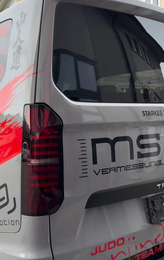 Ein weißer Van mit roten Designs an der Seite und hinten. Die Rückseite trägt die Aufschrift 'ms VERMESSUNG' und 'STARKES'. Das Nummernschild zeigt 'TF'.