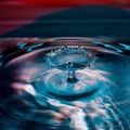 Ein Wassertropfen spritzt in einen Pool und erzeugt Wellen und Reflexionen. Das Wasser ist blau und klar, mit einem Lichtreflex auf der Oberfläche.