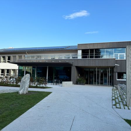 Der Eingang zu einem Gebäude mit Solarpanelen auf dem Dach, Glasfenstern und einem Gehweg, der zum Eingang führt. Ein großer Stein steht auf dem Rasen davor.