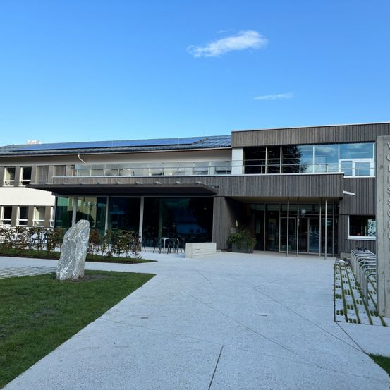 Der Eingang zu einem Gebäude mit Solarpanelen auf dem Dach, Glasfenstern und einem Gehweg, der zum Eingang führt. Ein großer Stein steht auf dem Rasen davor.