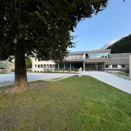 Ein modernes Gebäude mit Solarmodulen auf dem Dach steht in einem gepflegten Grasbereich. Ein Baum steht im Vordergrund.