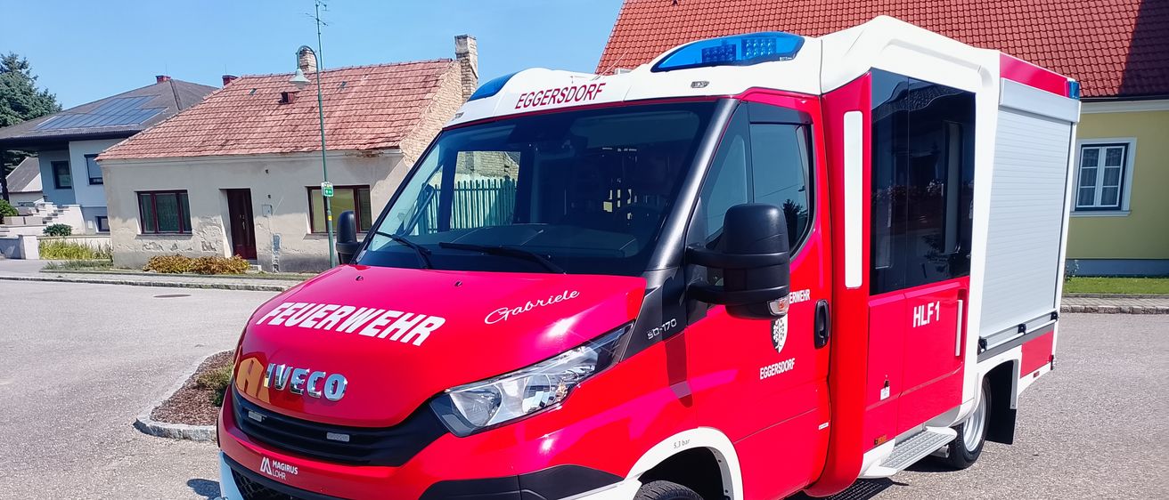 Ein roter und weißer Iveco-Feuerwehrwagen steht vor einem Haus mit rotem Dach. Der Wagen hat die Aufschrift Feuerwehr Eggersdoof an der Seite und ein Nummernschild mit dem Text FWI D313 MI.