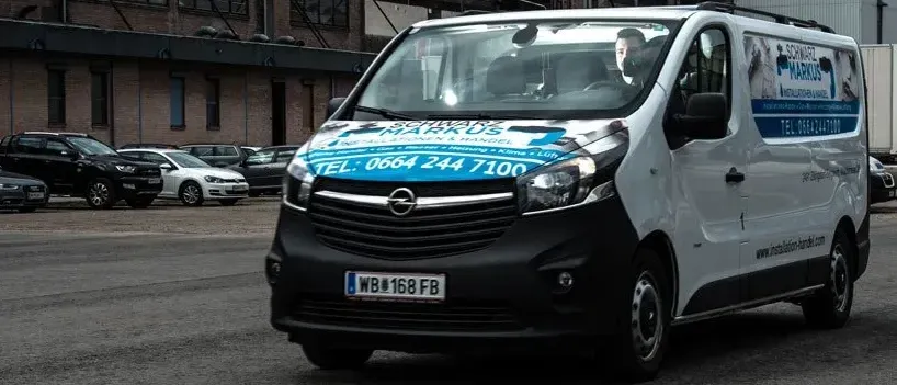 Ein weißer Lieferwagen mit blauer Werbung fährt an einem großen Backsteingebäude vorbei. Eine Person sitzt auf dem Fahrersitz.
