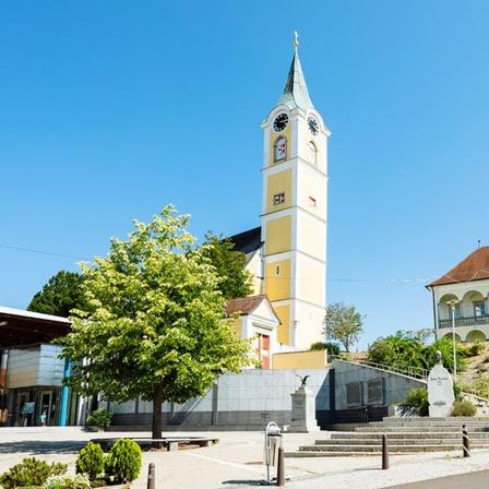 Ein hoher Uhrturm mit einer spitzen Spitze steht vor einem Gebäude. Bäume, Pflanzen und Büsche umgeben das Gebiet. Eine Statue steht auf den Stufen, die zum Turm führen.