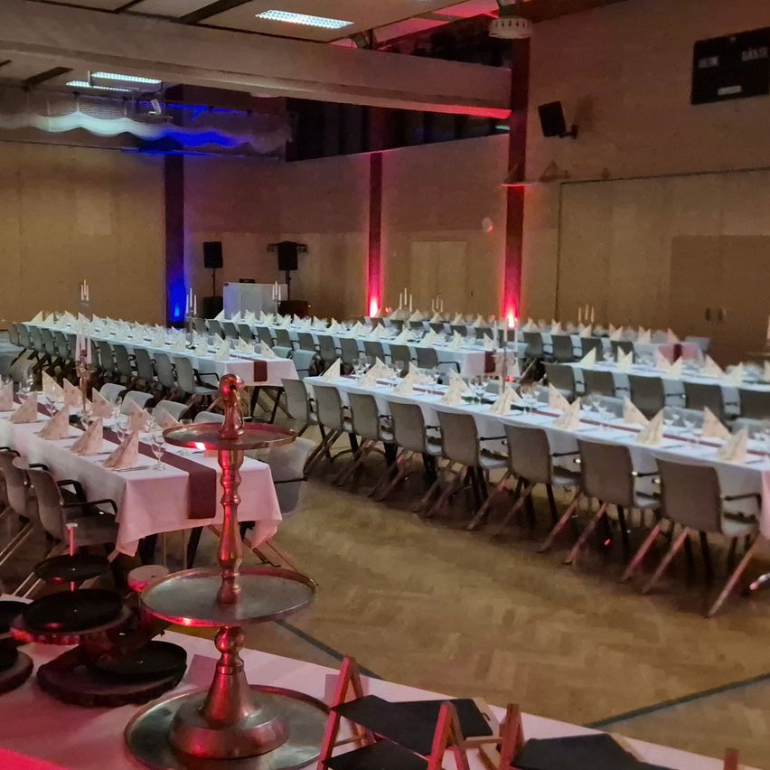 Ein Bankettsaal ist für eine Veranstaltung vorbereitet mit Tischen und Stühlen in Reihen angeordnet. Jeder Tisch hat eine weiße Tischdecke, ein Tischzentrum und gefaltete Servietten. Rote und blaue Lichter dekorieren den Raum.