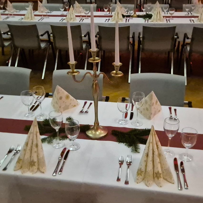 Ein Tisch ist für ein Abendessen mit roter Tischdecke, Weingläsern, gefalteten Servietten, Gabeln, Messern und einem Kerzenständer aus Messing mit Kerzen gedeckt. Dahinter sind weitere Tische gedeckt, und der Raum ist mit Stühlen gefüllt.