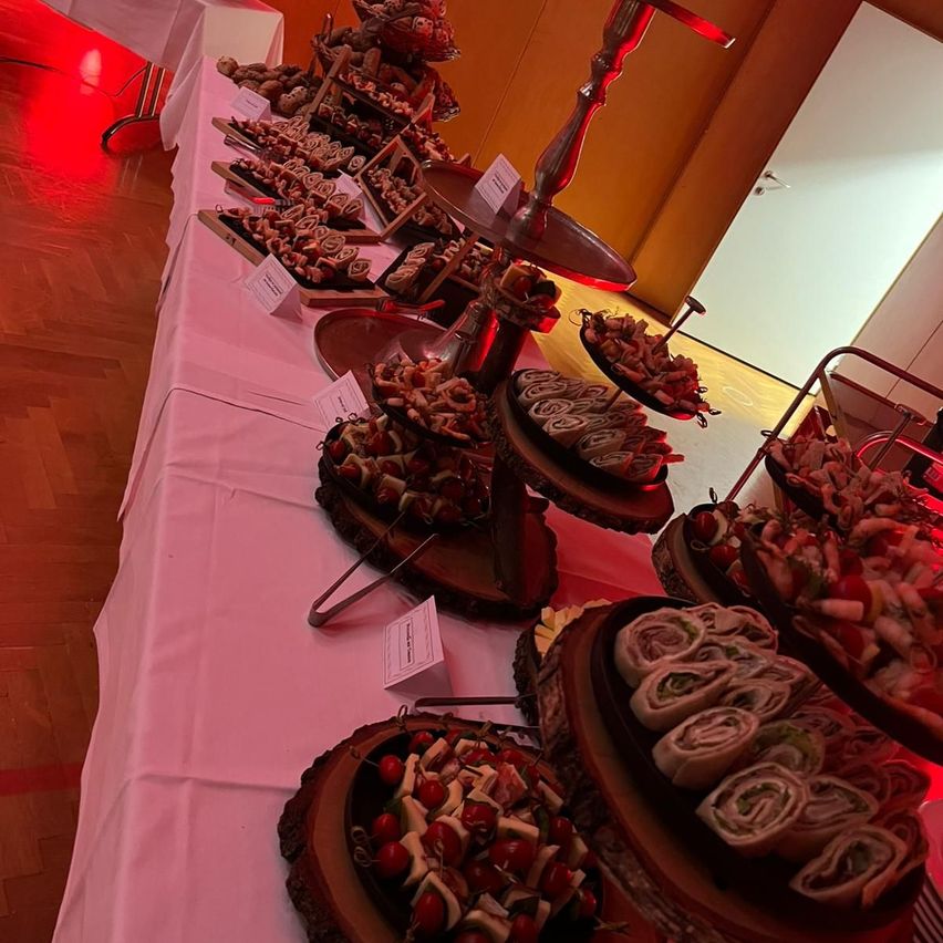 Ein langer Tisch, der mit einer rosa Tischdecke bedeckt ist, hält verschiedene Essensplatten, darunter Sandwiches, Brötchen und Spieße. Die Umgebung ist durch rotes Licht beleuchtet.