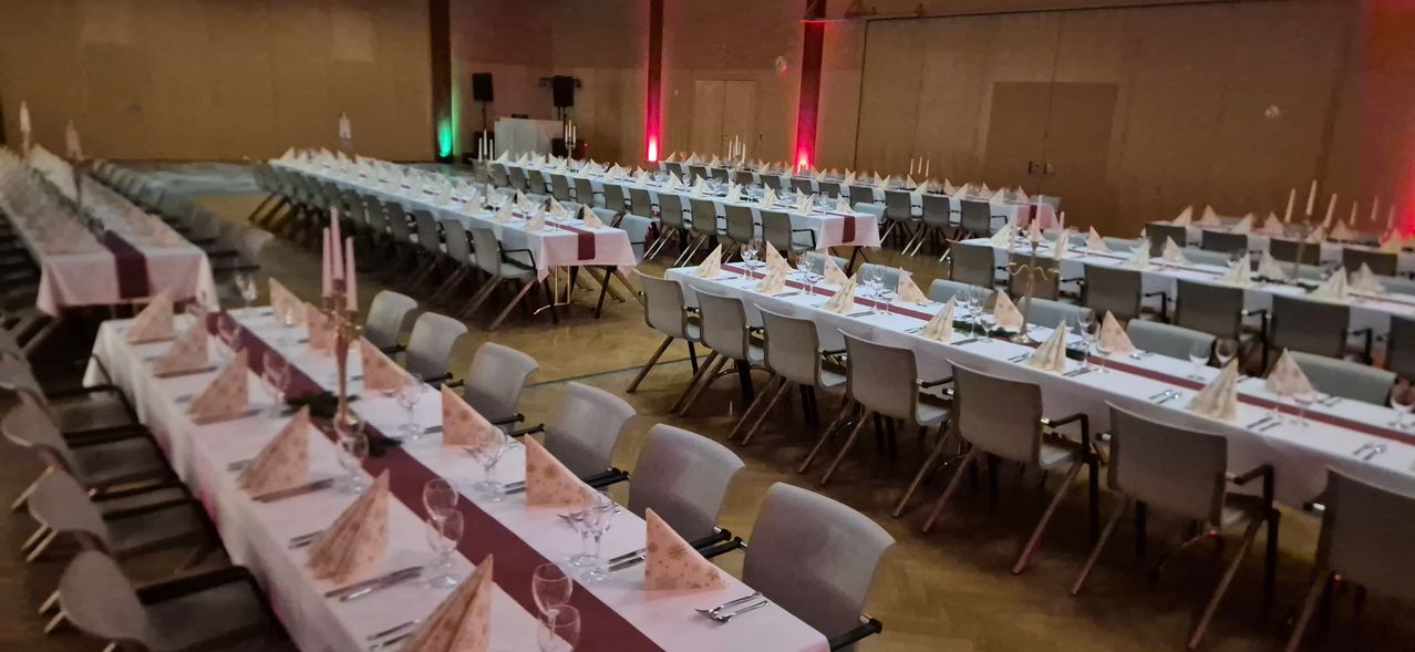 Ein Bankettsaal mit mehreren langen Tischen für eine formelle Veranstaltung, geschmückt mit weißen Tischdecken, Servietten, Weingläsern und Platzeinstellungen. Rote Beleuchtung betont den Raum.