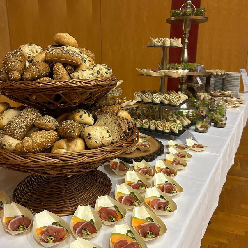 Ein langer Tisch zeigt eine Vielzahl von Speisen. Es gibt Körbe mit Brot, Sandwiches und Prosciutto in kleinen Papierbooten. Es gibt auch einen mehrstöckigen Ständer mit Mini-Sandwiches und Salaten.