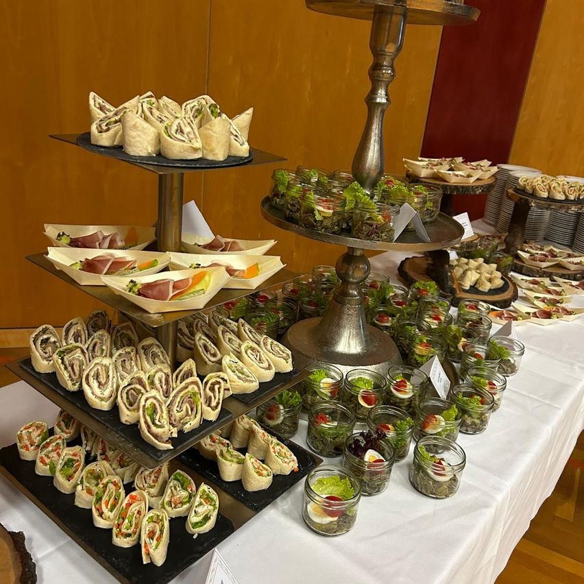 Ein Buffettisch mit mehreren geschichteten Tabletts, die Sandwiches und Salate halten, sowie kleinen Gläsern mit Salat, ist auf einer weißen Tischdecke ausgestellt. Der Hintergrund zeigt eine Holzwand.