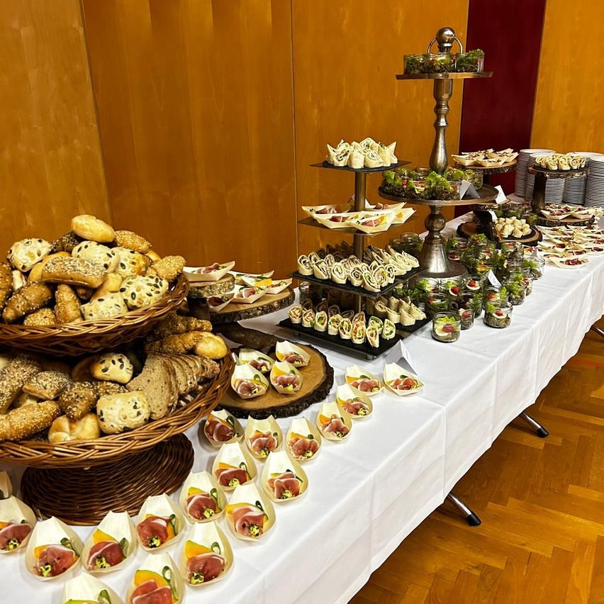 Ein Tisch mit einer weißen Tischdecke hat eine Vielzahl von Speisen wie Sandwiches, Wraps und Brot auf Tabletts. Ein Stapel Teller ist rechts platziert.