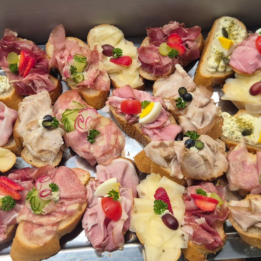 A tray of gourmet sandwiches, each topped with a variety of meats, cheese, and fresh vegetables. The bread slices are accompanied by colorful fruits and olives.
