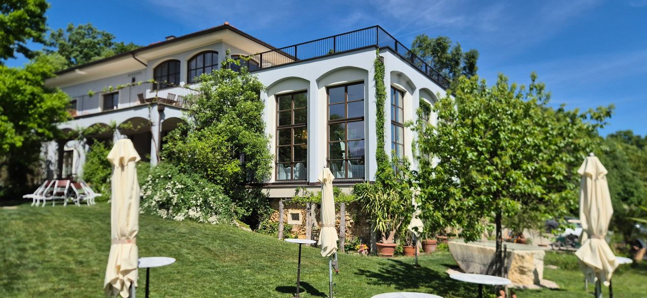A two-story house with a balcony and large windows, surrounded by lush greenery and a grassy lawn. Potted plants and a folded umbrella add to the scene.