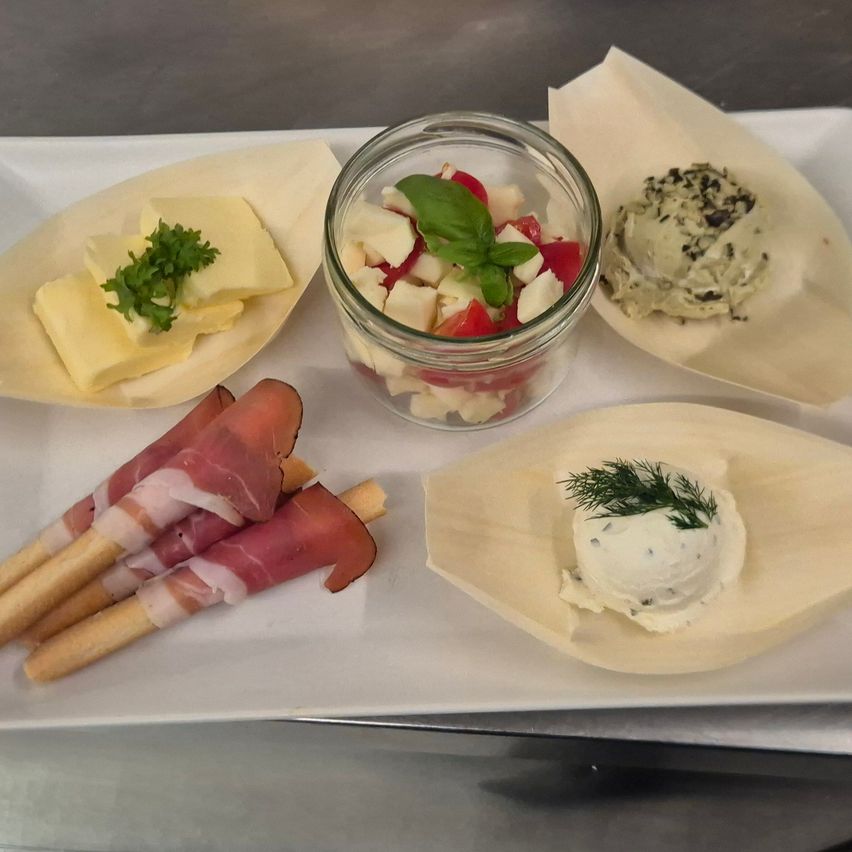 A white plate with various foods on it, including slices of cheese, a bowl of salad, and a bowl of cream cheese. There are also skewers of meat and a small piece of meat.