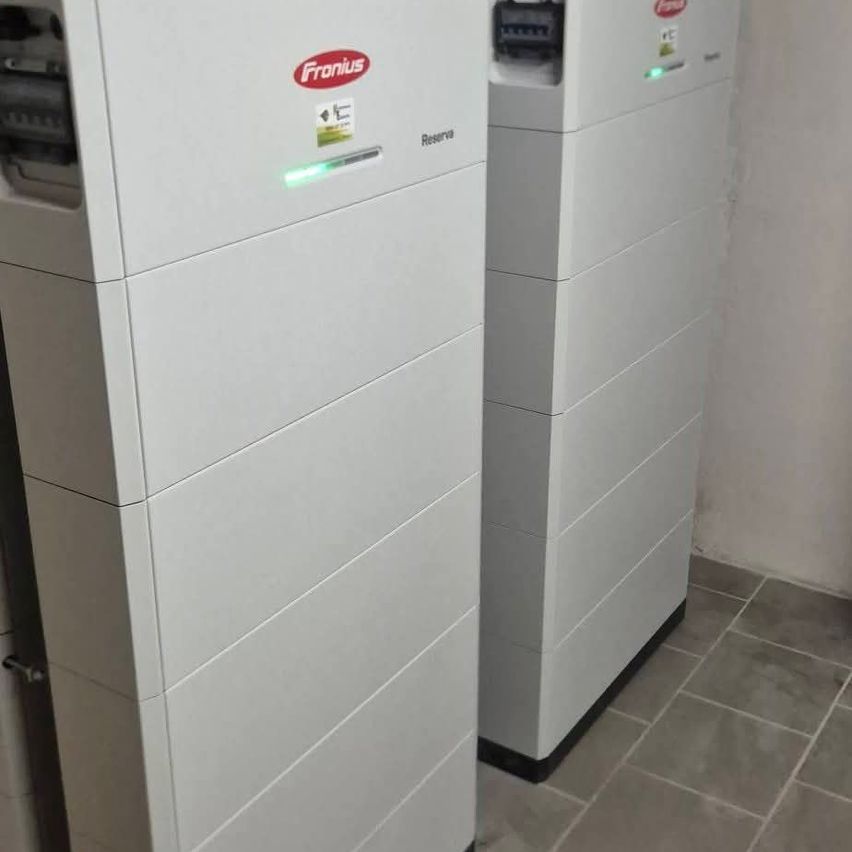 Two white industrial devices are stacked. The left device has a red 'FRONIUS' logo and a green light. The right device has a red sticker. Both sit on tiled floor.