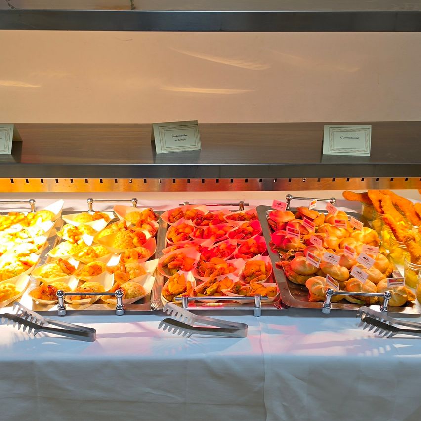 A buffet table with various pastries, including fried dough, croissants, and bread rolls, displayed on trays with labels and tongs for serving.