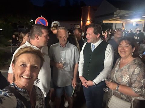 Eine Gruppe von Menschen steht bei Nacht draußen und lächelt, während sie Weingläser halten. Im Hintergrund sind Gebäude und Bäume zu sehen.