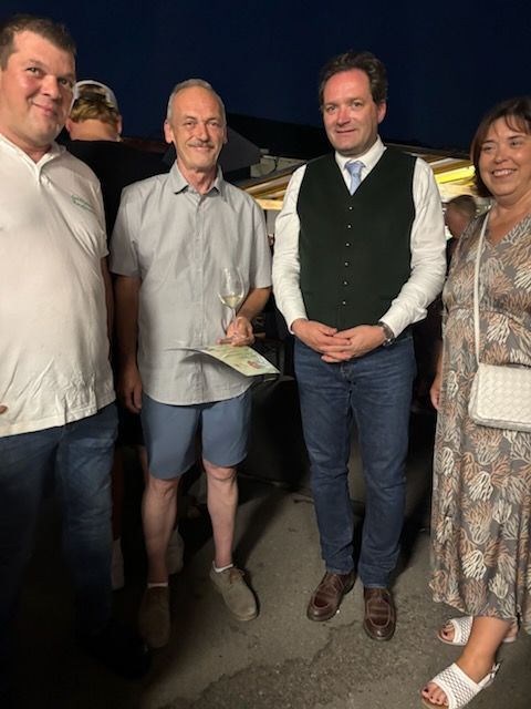Vier Menschen stehen lächelnd für ein Foto zusammen. Ein Mann hält ein Weinglas und Papier. Ein anderer Mann trägt eine Weste. Eine Frau trägt eine weiße Tasche.
