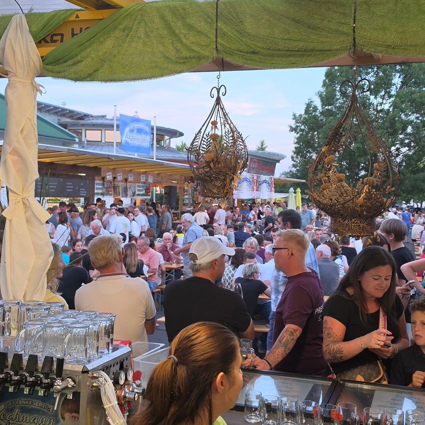Ein überfülltes Biergarten im Freien mit Menschen, die an Tischen sitzen, trinken und den Abend genießen. Es gibt dekorative hängende Laternen und eine Bar mit Gläsern und einem Zapfhahn.