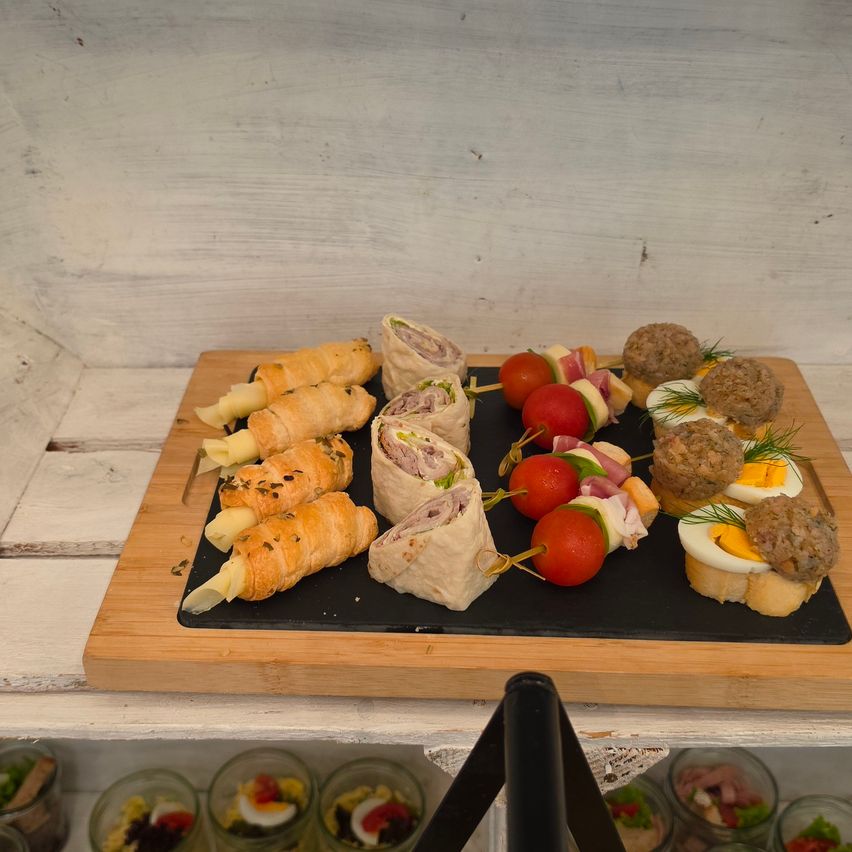 Ein Holztablett zeigt verschiedene Lebensmittel wie Sandwiches, Fleischbällchen und Spieße mit Tomaten und Eiern. Der Hintergrund ist eine weiße Wand.