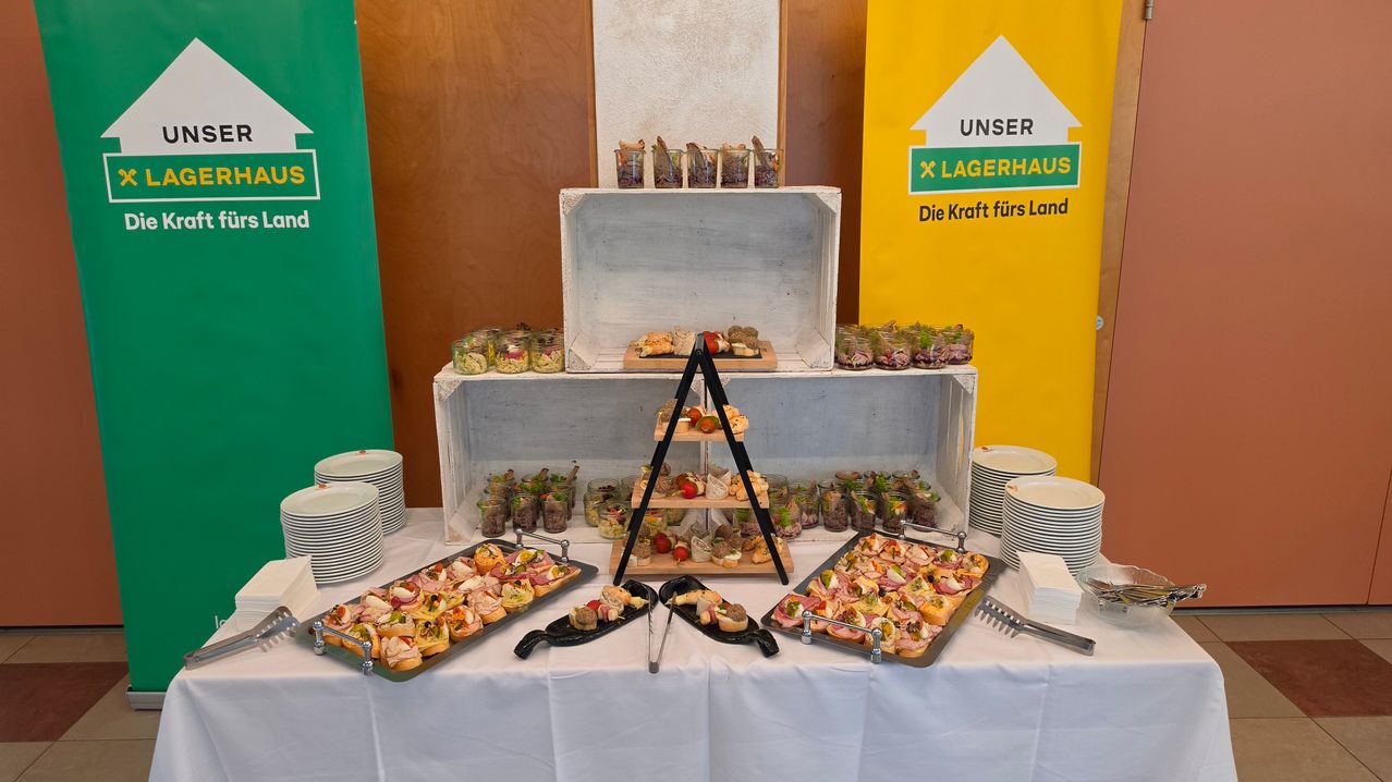 Ein Buffettisch mit verschiedenen Vorspeisen auf Tabletts, darunter Sandwiches und Salate, auf einer weißen Tischdecke angeordnet. Dahinter befindet sich eine Holzkiste mit weiteren Lebensmitteln, und ein gelber Banner mit dem Text 'UNSER LAGERAUS' ist sichtbar.