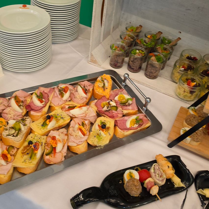 Ein Tisch mit verschiedenen Sandwiches, Fleisch und Salaten. Ein Tablett mit Sandwiches mit Fleisch und Gemüse steht auf einem silbernen Tablett. Kleine Gläser mit Salaten und Fleisch stehen auf einem weißen Regal.