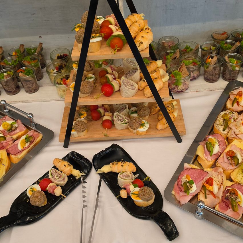 Ein dreistufiges Tablett gefüllt mit verschiedenen Sandwiches und Vorspeisen, einschließlich Brötchen, Fleischscheiben und Gemüse, mit zusätzlichen Gerichten in Gläsern und auf Tabletts in der Nähe.