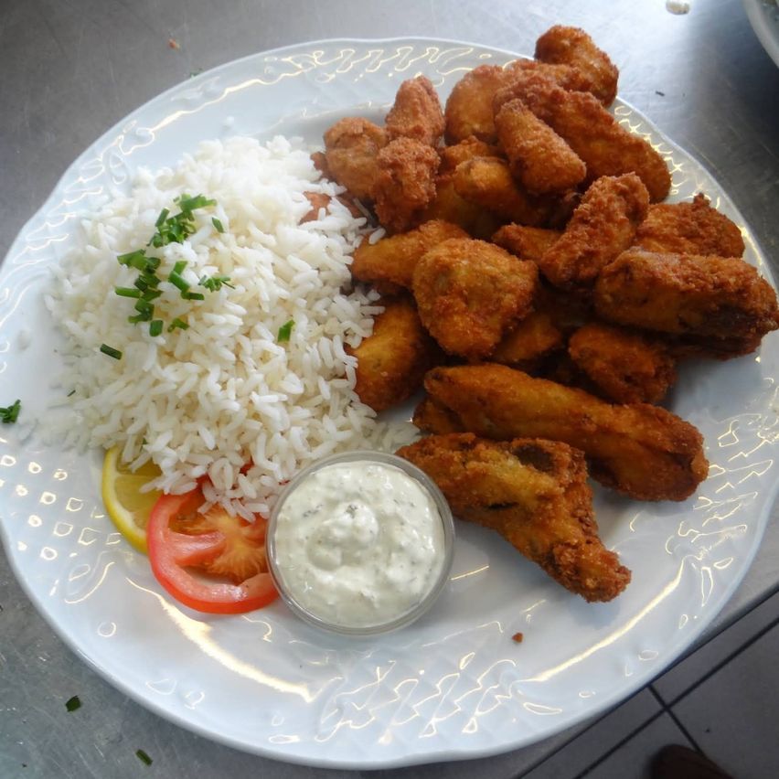 Ein weißer Teller mit gebratenem Hühnchen, Reis und Dip. Das Hühnchen ist goldbraun und der Reis ist locker. Es gibt auch geschnittene Tomaten und eine Zitronenscheibe.
