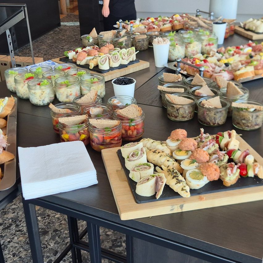 A table full of various sandwiches, meats, and vegetable platters. The food is served in jars, bowls, and plates. The table is surrounded by napkins and a black cutting board.