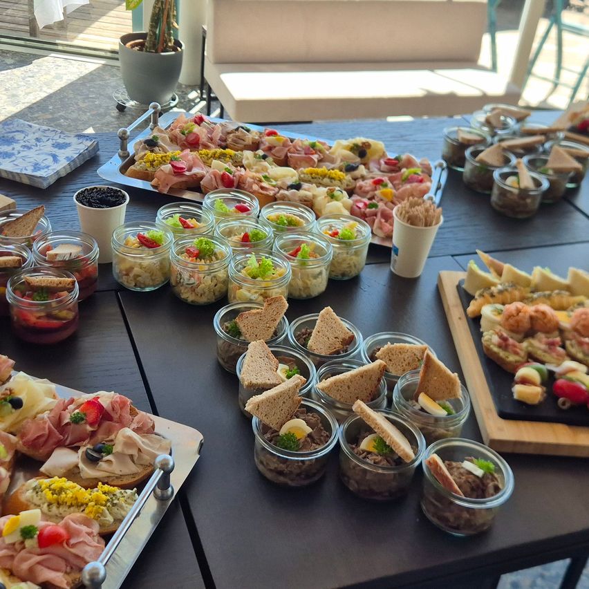 A wooden table with various sandwiches and meat in jars. On the right, a plate with meats and olives. A white sofa is in the background.