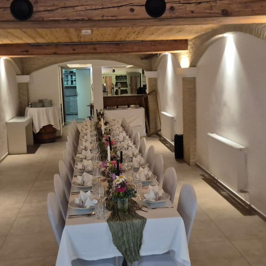 A long table with white cloth and plates set for a meal is in a room with wooden beams and white walls.