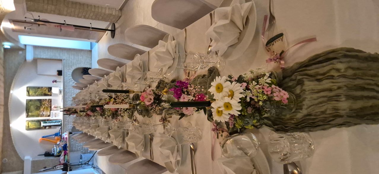 A long table is beautifully set with white napkins, wine glasses, and floral centerpieces. The table is covered with a white tablecloth, and the flowers include daisies, pink and purple flowers, and green leaves.