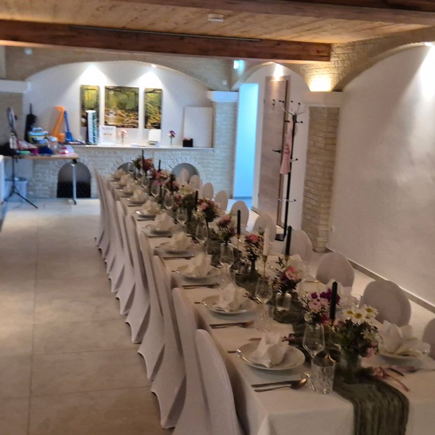 A long dining table set with flowers, plates, and cutlery. The room has a brick wall, a wooden ceiling, and a back area with a table and artworks.