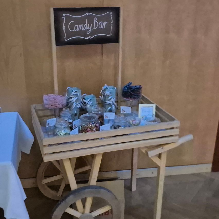 A wooden cart with a sign reading 'Candy Bar', filled with various candies in glass jars. The cart is placed against a wall, next to a white tablecloth.
