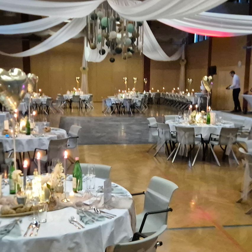 A hall decorated for an event, featuring multiple round tables with white cloths, candles, and decorative items. A chandelier hangs from the ceiling, and a man stands near the stage.