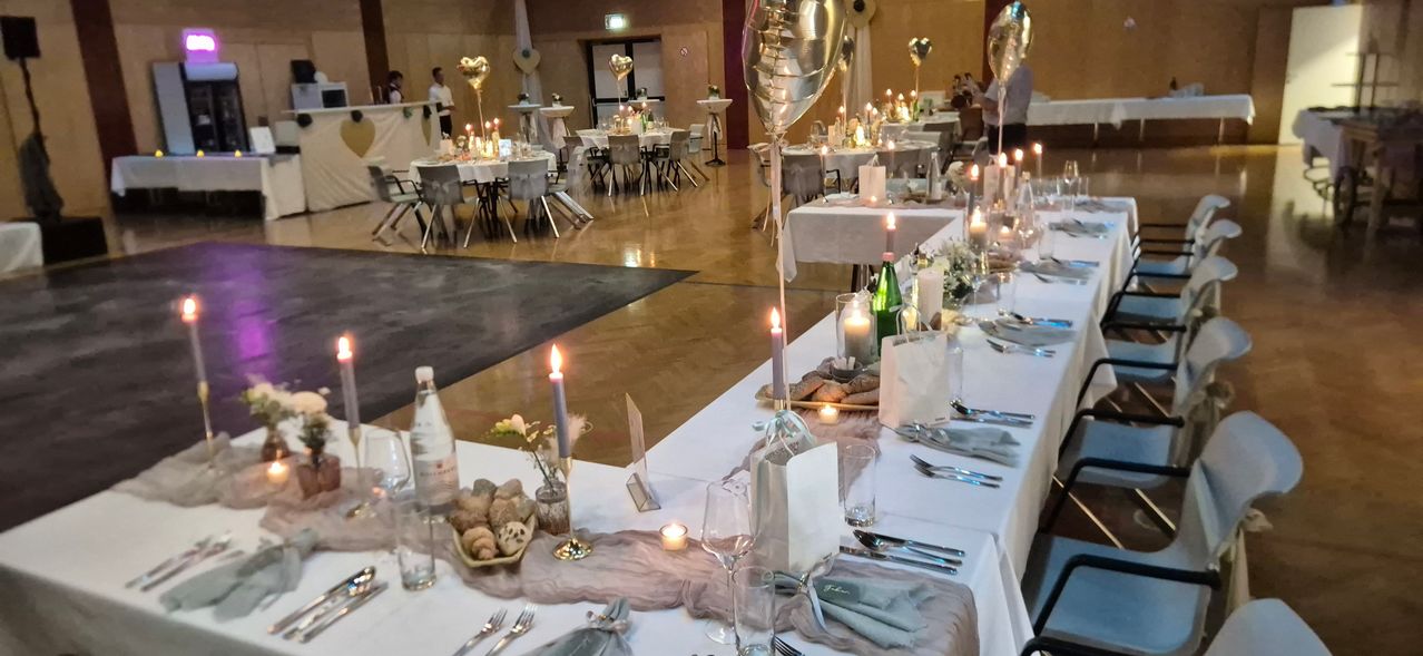 A large dining hall with multiple tables, each with candles, balloons, and place settings. The tables are covered with white tablecloths.