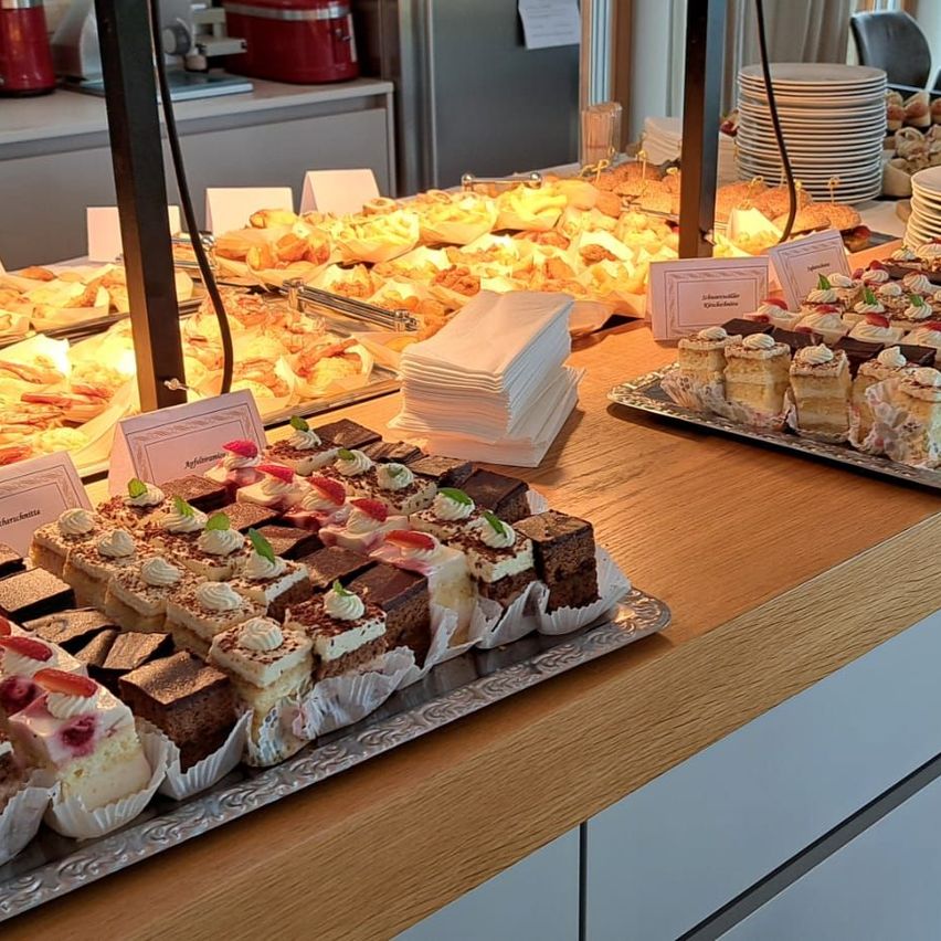 Eine Buffet-Tabelle ist mit einer Vielzahl von Desserts gefüllt, einschließlich Kuchen, Gebäck und Törtchen. Der Tisch ist mit einer hölzernen Tischdecke bedeckt.
