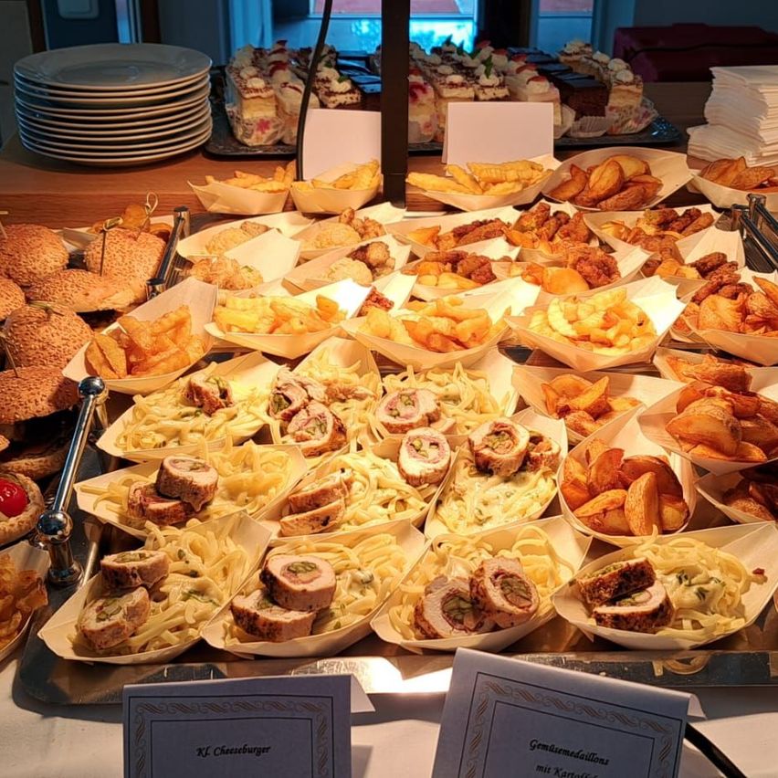Ein Buffettisch mit verschiedenen Speisen, darunter Sandwiches, Pasta und Pommes Frites, alles ordentlich in Papierbehältern angeordnet. Ein Schild mit der Aufschrift 'Xf Cheeseburger' ist auf dem Tisch platziert.