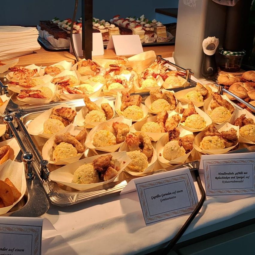 Ein Buffettisch mit verschiedenen Mini-Gerichten und Gebäck, darunter panierte frittierte Speisen, Mini-Quiches und Desserts. Ein weißes Schild mit der Aufschrift 'Geräucherte Garnelen auf einem Gemüsebett'.