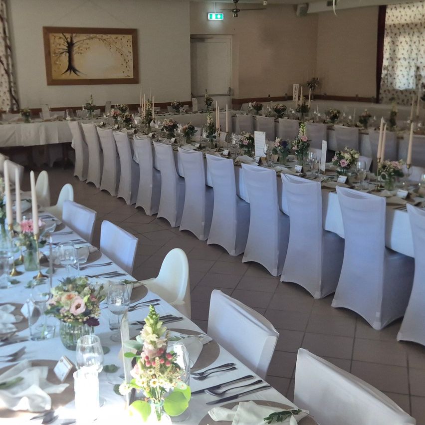 A large banquet hall with long tables set for an event, each with white chairs, floral centerpieces, and candles. The room has white walls, a picture frame, and a ceiling fan.