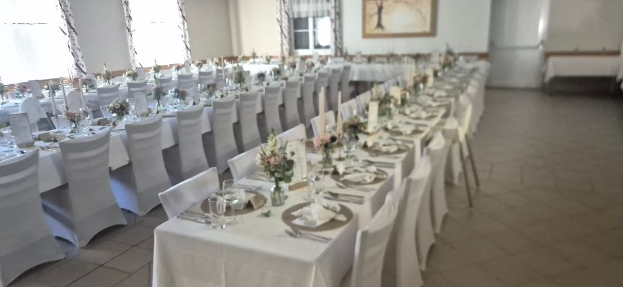 A long banquet table set for a formal event with white tablecloths, floral centerpieces, and white chairs. The table is adorned with plates, silverware, and wine glasses.
