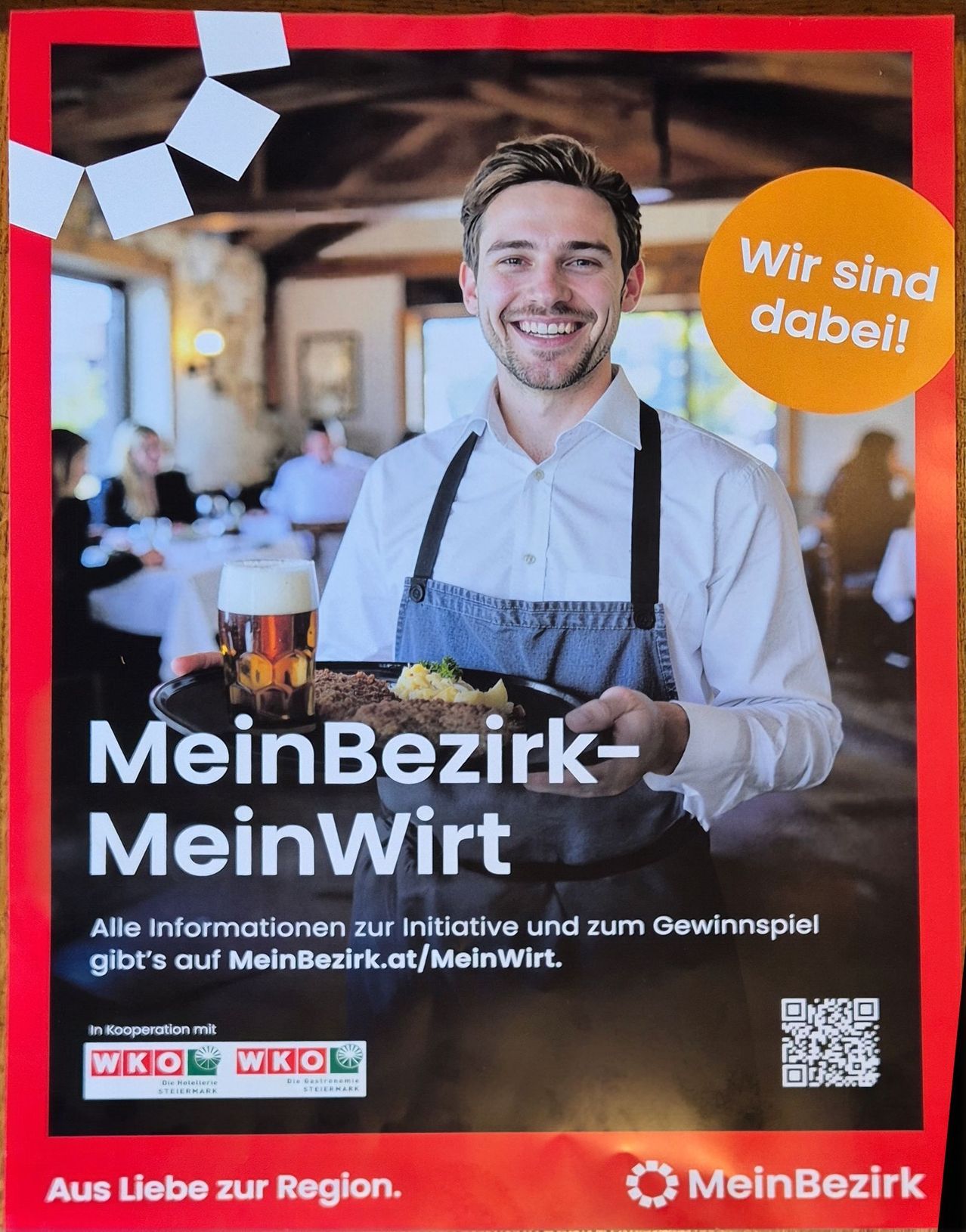 A smiling man in a restaurant holding a tray with food and a glass of beer. The poster says 'MeinBezirk-MeinWirt' and 'Wir sind dabei!' with a QR code.