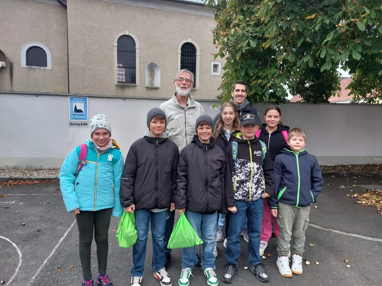 Eine Gruppe von Kindern und zwei Erwachsenen steht vor einem Gebäude. Sie alle tragen Jacken und halten Taschen. Das Gebäude hat Bogenfenster. Ein Schild an der Wand steht für 'Messe'. Der Baum im Hintergrund ist grün.