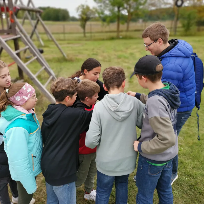 Eine Gruppe von Kindern und ein erwachsener Mann versammeln sich auf einer Wiese und bilden einen Kreis. Der erwachsene Mann trägt eine Brille und eine blaue Jacke.