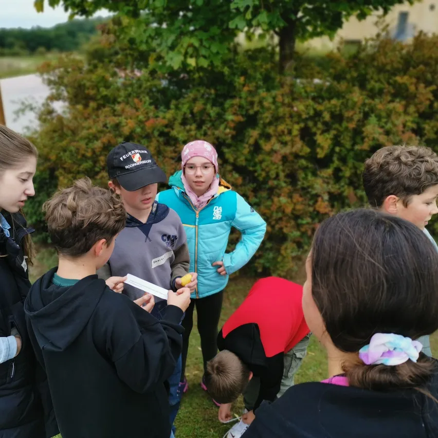 Eine Gruppe von Kindern steht auf einer Rasenfläche, wahrscheinlich spielen sie ein Spiel. Einige schauen nach unten, während andere den Jungen vor ihnen betrachten. Sie sind von einem Busch und Bäumen umgeben.