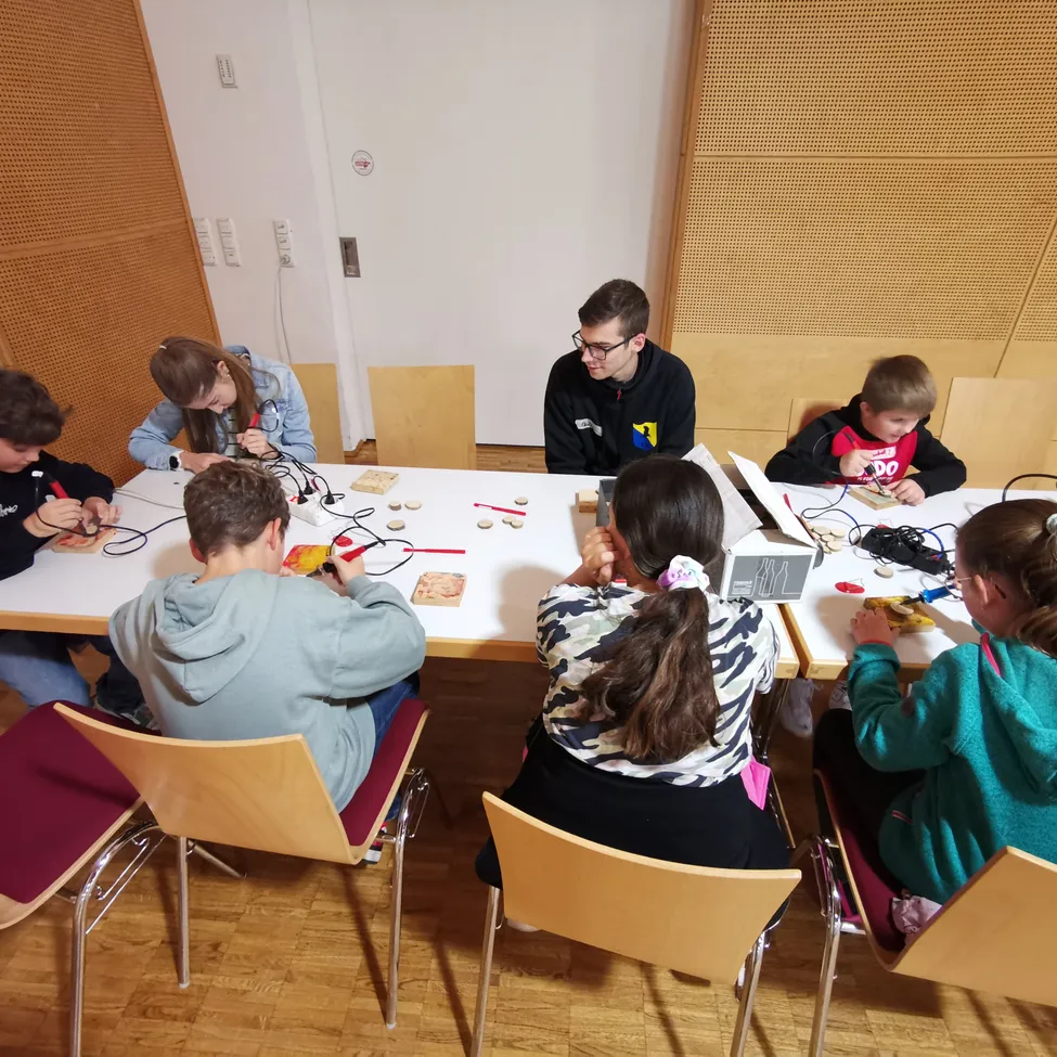 Eine Gruppe von Kindern und einem Mann sitzen um einen Tisch herum und beschäftigen sich mit einer Aktivität mit elektronischen Komponenten. Der Mann trägt eine Brille und eine schwarze Jacke. Die Kinder, die Jacken tragen, scheinen konzentriert auf ihre Aufgaben. Der Tisch ist mit verschiedenen elektronischen Teilen und Werkzeugen bestückt.